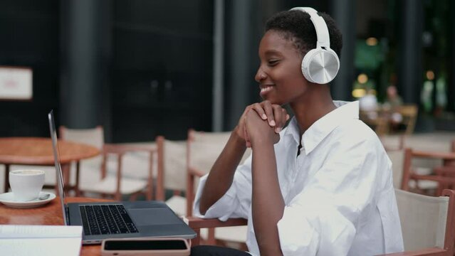 focused young Black woman in headphones participating in online English learning class. Seated at outdoor caf&eacute;, female interacts with her laptop computer, absorbed in lesson. educational atmosphere
