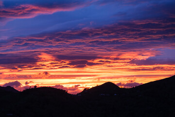 A vibrant sunset casts a warm glow over the sky, with fluffy clouds drifting above towering mountains in the background. The suns rays create a stunning display of colors on the horizon.