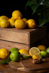 Closeup of table with wooden box with lemons, limes, board, green leaves and sugar, on wooden table, black background, vertical, with copy space