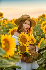 A girl in a hat on a beautiful field of sunflowers against the sky in the evening light of a summer sunset. Sunbeams through the flower field. Natural background.