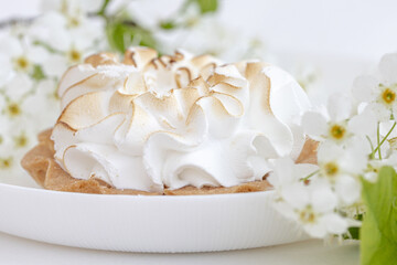 Delicious tartlet with meringue on a white plate, a branch of blooming cherry tree. Sweet treat in spring or summer, copy space