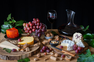 Top view of rustic table with pieces of cheese, red grapes, glass and decanter of red wine, nuts and bread, black background, horizontal