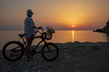 A young woman with a bicycle enjoys the sunset by the sea. 