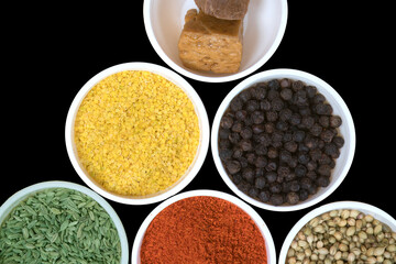 close-up view of small white bowls filled with Asafoetida, Yellow Split Mustard Seeds, Black Pepper, Fennel Seeds, chili pepper powder, Coriander Seeds, Isolated on Black Background