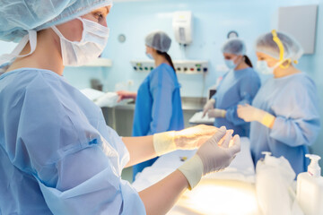 Caucasian female surgeon preparing for operation in a busy operating room with medical team. All team members are wearing surgical gowns, masks, and gloves