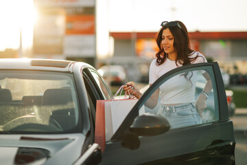 Young trendy woman with shopping paper bags entering car on parking.