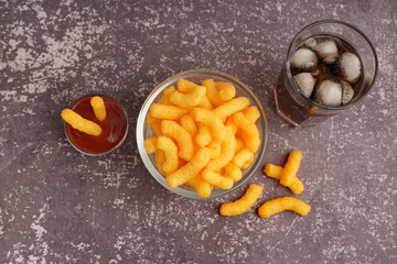 Bowl with tasty salty corn sticks, glass of cola and ketchup on grunge background