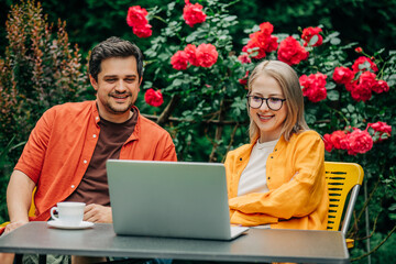 man and woman sitting together at outdoor table in a garden setting with roses, surrounded by vibrant red flowers and lush greenery. Both are engaged with a laptop