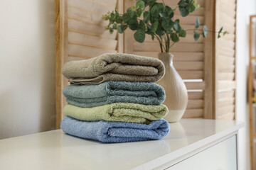 Stack of clean towels and vase with green branches on chest of drawers  near folding screen, closeup