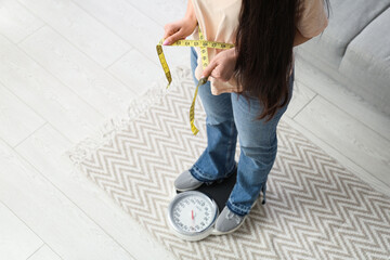 Beautiful young sporty woman with measuring tape and scales at home, closeup. Weight loss concept