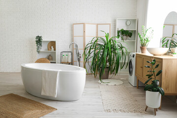 Interior of stylish bathroom with bathtub, chest of drawers, houseplants and mirror