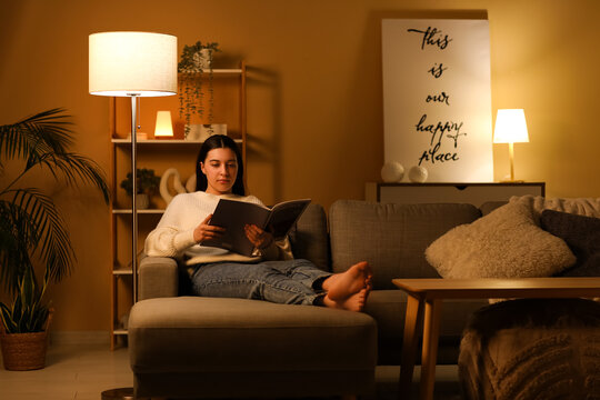 Young woman reading magazine and resting on cozy sofa in living room at night