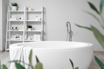 Interior of stylish bathroom with bathtub, shelving unit and olive tree leaves