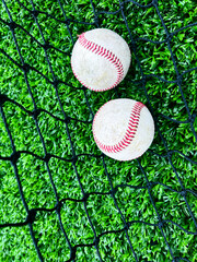 Two Baseballs on Green Turf. Two baseballs inside of a batting cage. The two balls rest upon the black sports net and artificial green turf.