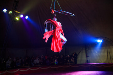 Professional aerial hoop woman gymnast circus performance at red arena with dark background backlit with colored concert lights, performing gymnastic elements