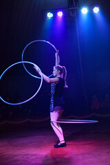 Young slim attractive circus gymnast woman performing live show with hula hoops on red arena with blue backlight, audience is excited