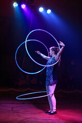 Young slim attractive circus gymnast woman performing live show with hula hoops on red arena with blue backlight, audience is excited