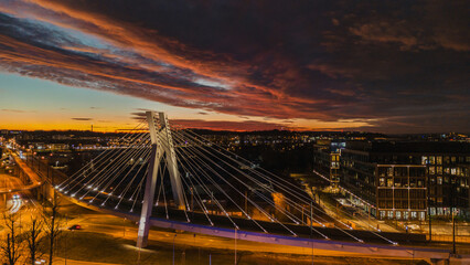 Panoramic night perspective scene with bridge and roads in lights, Krakow, Poland. High panorama view of bend of bridge hanging on steel cables on concrete structure, dramatic red sunset clouds in sky