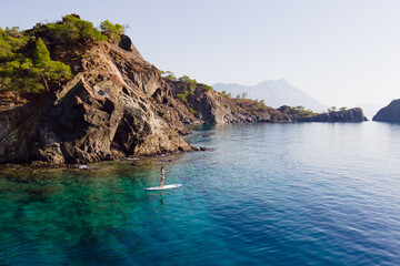 Aerial view of woman paddling a stand-up paddleboard or SUP board on a calm sea along rocky shore