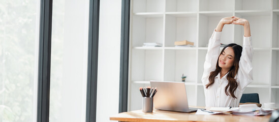 A young woman stretches at her desk in a modern office, taking a relaxing break from work. The...