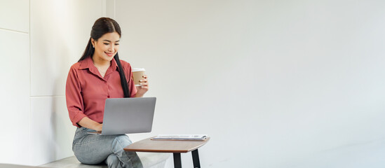 A woman is sitting on a stool with a laptop in front of her. She is drinking coffee while working...