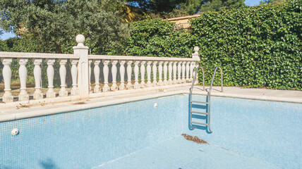 Blue tiled walls and steps of an empty dry swimming pool. Background of green leaves. Vacation low season. Square pattern of ceramic tiles. Roman columns railing for pool fence. Colonial style fence