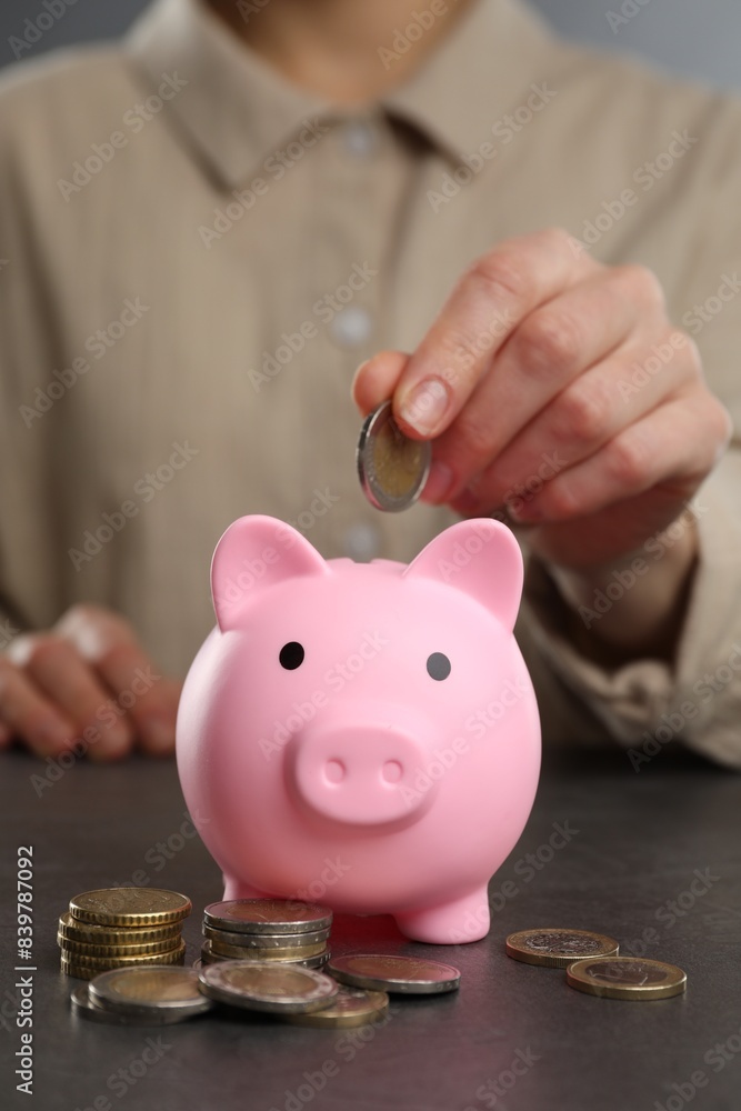 Wall mural Woman putting coin into pink piggy bank at black table, closeup