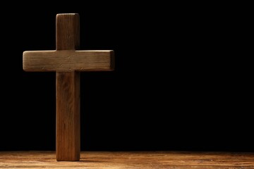Cross on wooden table against black background, space for text. Religion of Christianity