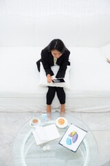 Young asian businesswoman sitting on sofa using a digital tablet with work documents while coffee...