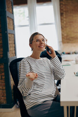 Young smiling female entrepreneur talking by phone with client while sitting in modern office 