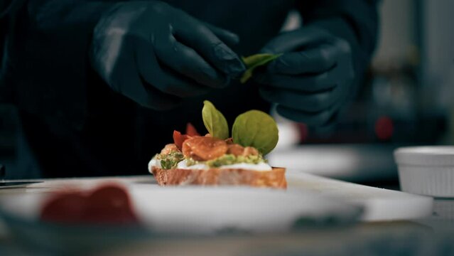 close up in a professional kitchen a chef in a black jacket decorates toast with salmon with leaves