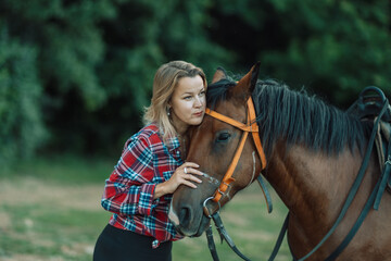 Obraz premium Happy blonde with horse in forest. Woman and a horse walking through the field during the day. Dressed in a plaid shirt and black leggings.