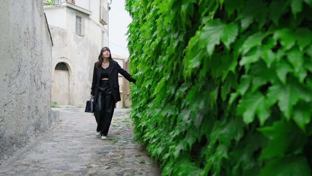 Woman Dressed In Black Walks Touching Ivy In A City Alley Of Gerace