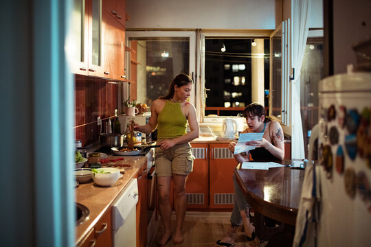 Lesbian couple cooking and reviewing documents in their apartment kitchen at night - Powered by Adobe