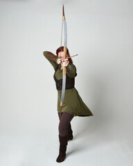 Full length portrait of red haired female model wearing green medieval fantasy costume, leather armour.
Holding archery bow and arrow weapon, standing action pose isolated on white studio background.