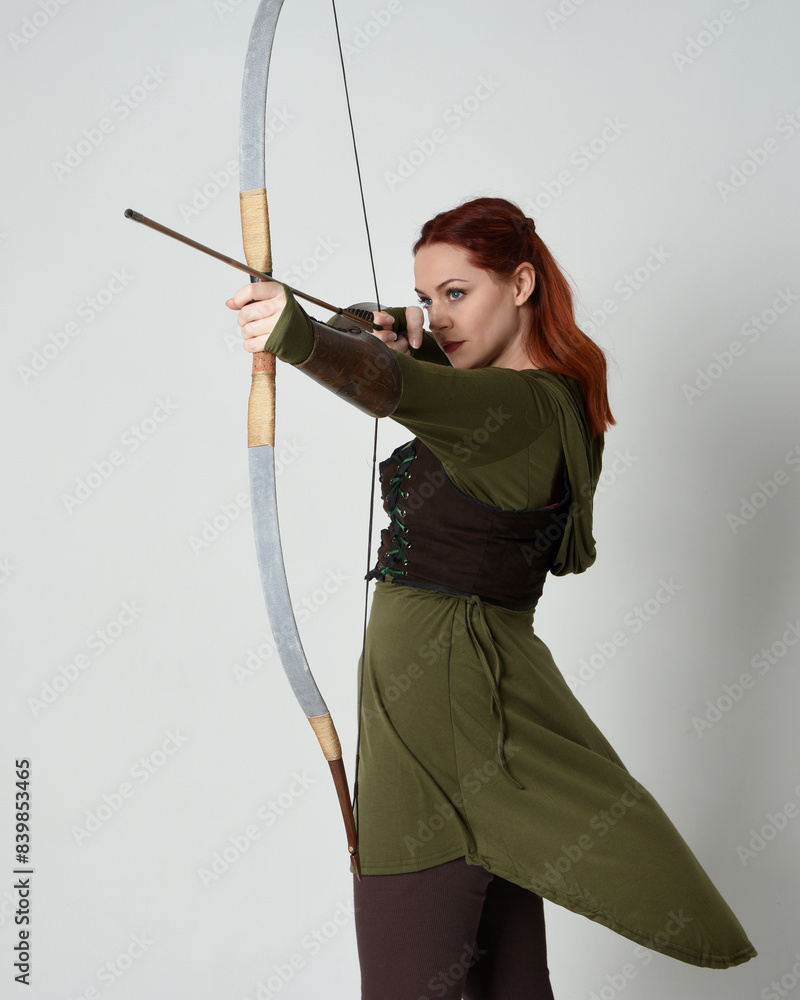 Poster close up portrait of red haired female model wearing green medieval fantasy costume, leather armour.
Holding archery bow and arrow weapon, standing action pose isolated on white studio background.