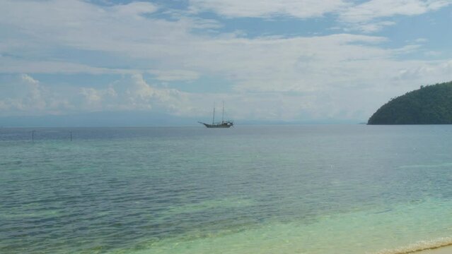 Traditional sailing boat floats in a distance on the turquoise waters of Raja Ampat, Indonesia. The serene sea creates tranquil scene of untouched beauty in tropical paradise. Static slow motion shot.