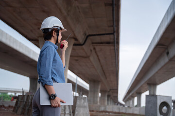 Asian female engineer with a laptop, managing the construction project under an overpass, ensuring...
