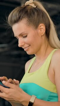 A pretty young blonde woman is walking on a treadmill in the gym and is typing a message on her mobile phone. She is smiling. Vertical shot.