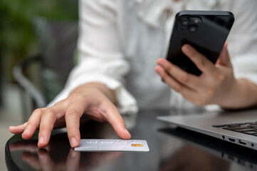 A woman is touching a credit card on a table, looking at her credit card number and using her phone.