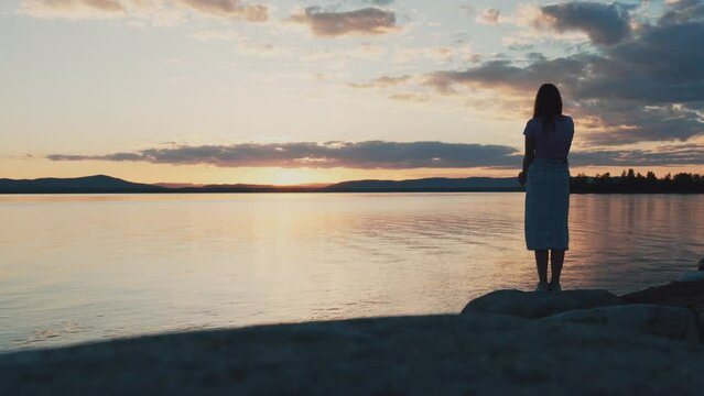 The silhouette of a lonely young girl standing on the shore of a lake at sunset. The girl admires the beauty of nature. The harmony of nature and man.
