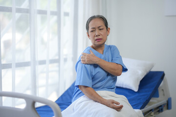 An elderly woman in a hospital room experiencing shoulder pain. She sits on a bed, wearing blue...