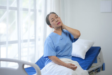 Woman in hospital gown sitting on a bed, holding her neck in discomfort, in a bright hospital room with white curtains.