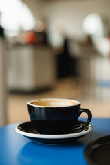 A black cup of coffee on a blue table against a blurred and unrecognizable background of a coffee shop interior.