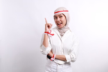 A happy Asian muslim woman wearing red white flag ribbon and pointing copy space on top of her, isolated by white background. Indonesia's independence day