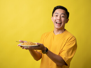 A young man in a yellow shirt is enthusiastically eating a plate of noodles with a fork against a...
