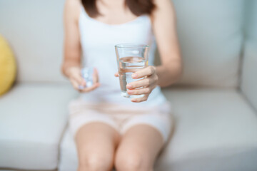 woman hand holding medicine painkiller pill and water glass on the sofa at home, taking for...