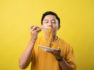 A young man in a yellow shirt is enthusiastically eating a plate of noodles with a fork against a...