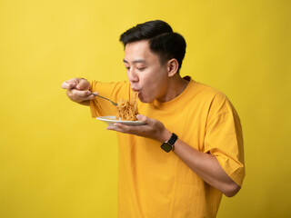 A young man in a yellow shirt is enthusiastically eating a plate of noodles with a fork against a...