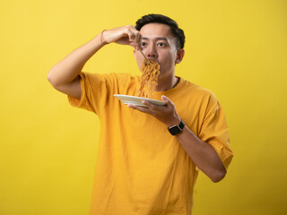 A young man in a yellow shirt is enthusiastically eating a plate of noodles with a fork against a...